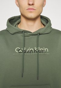 Felpa verde oliva di Calvin Klein con stampa del logo a contrasto, cordini con punte nere e un tessuto morbido e testurizzato.