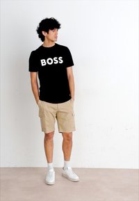 Melnas kokvilnas T-krekls ar baltiem burtiem "BOSS", kas apvienots ar khaki krāsas cargo šortiem un baltiem sporta apaviem. Vienkāršs dizains, ikdienišķs piegriezums.