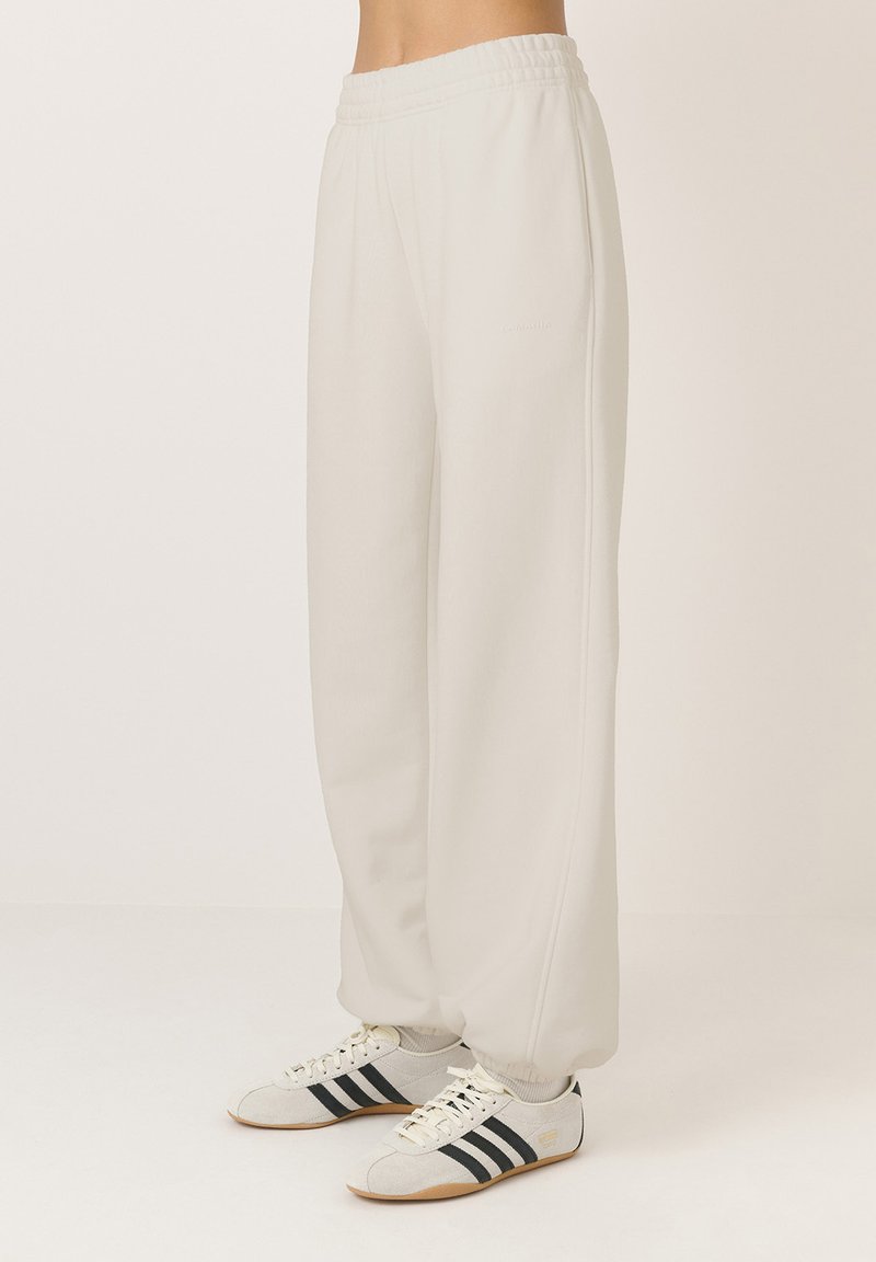 Világoskrém színű melegítőnadrág, elasztikus derékkal, széles lábakkal és puha textúrával; szürke sportcipővel párosítva, fekete csíkokkal.