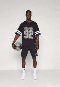 Must suurendatud t-särk tekstiga "CHAMPION 92" valgete triipudega. Mustad lühikesed püksid logoga; valged tossud; käes must-valge korvpall.