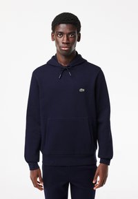 Felpa blu navy, realizzata in tessuto morbido, con cappuccio regolabile e tasca a marsupio frontale. Presenta un piccolo logo verde di Lacoste.