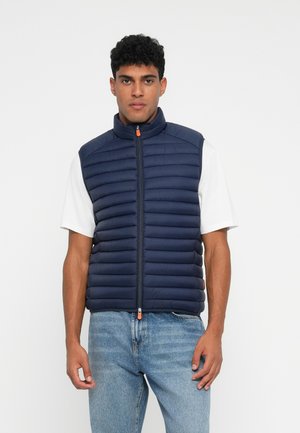 Navyblauwe puffer vest met horizontale stiksels, hoge kraag en een voorrits met een oranje trekstang, gedragen over een wit shirt.