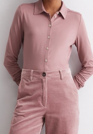 Person wearing a long-sleeve mauve button-up polo shirt tucked into high-waisted dusty pink corduroy pants.