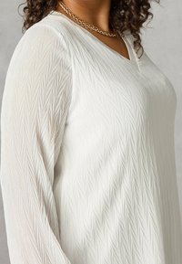 Donna che indossa un top bianco a maniche lunghe con una trama, abbinato a una collana di catena dorata, che mostra il torso superiore e i capelli ricci alla spalla.