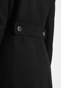 Svart kappa med ett slätt, strukturerat material, horisontell bältesdetalj och två knappar vid midjeområdet.