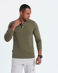 Man i olivgrön långärmad henley-tröja och ljusgrå cargobyxor som tittar ner, justerar halsringningen, och bär en svart armbandsklocka.