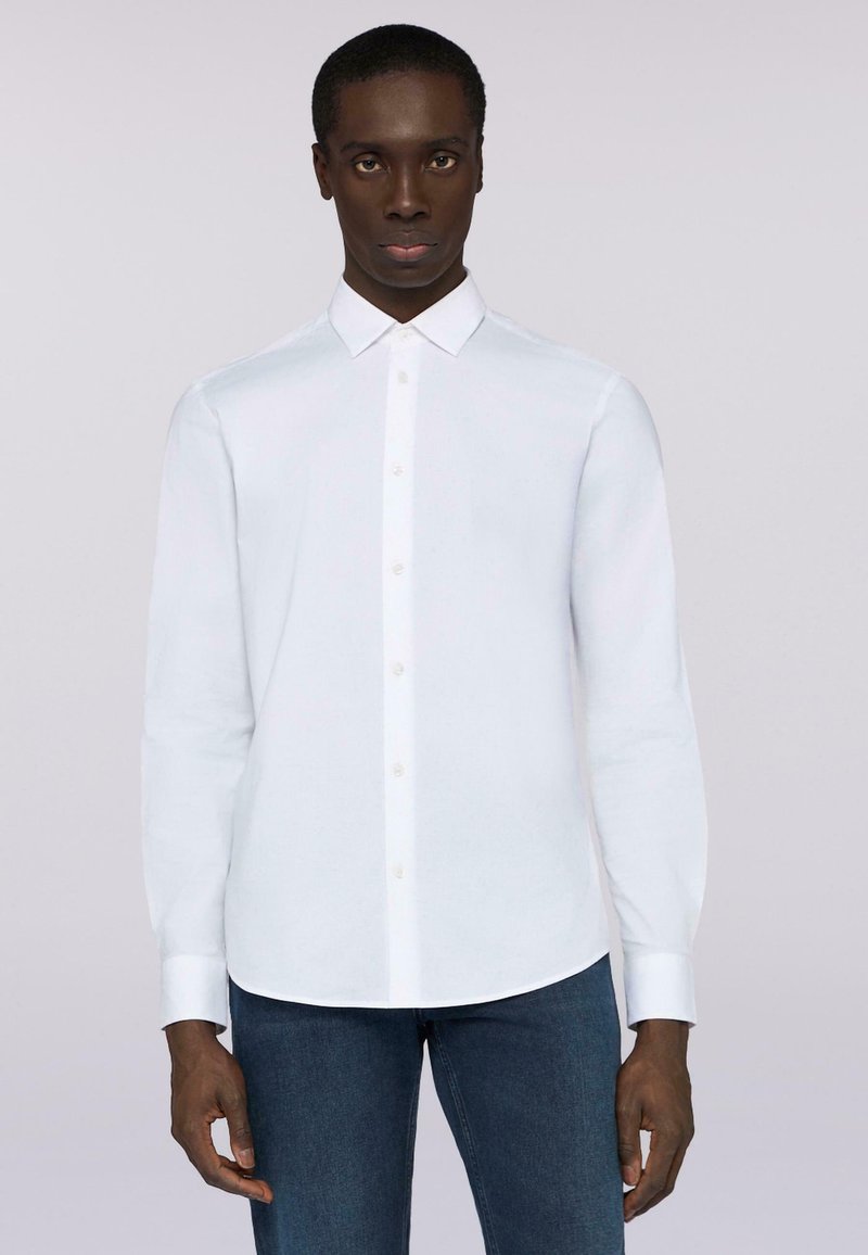 Biała koszula z długim rękawem i kołnierzykiem, gładka w dotyku, z zaokrąglonym dołem. Połączona z niebieskimi dżinsami.