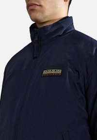 Mörkblå zip-up-jacka i lätt tyg; har en hög krage och en logotyp-patch på bröstet. Slät textur, avslappnad design.