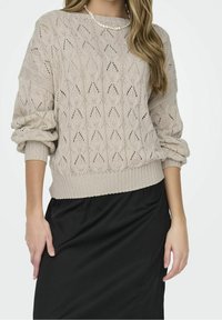 Beżowy sweter dziergany z wzorem w diamenty, o szerokich rękawach i ściągaczu u dołu. Nosi się go na czarnej spódnicy, ma okrągły dekolt.