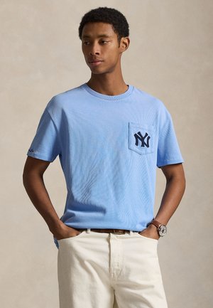 Világoskék, bordázott pamut póló kerek nyakkal és rövid ujjakkal; elöl zsebet és sötétkék NY logót tartalmaz. Krémszínű nadrággal viselve.