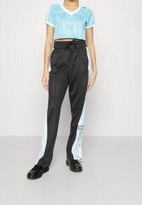 Person som bär en ljusblå kortärmad Adidas-tröja, svarta träningsbyxor med vita och blå sidostråkar, och svarta skor, står mot en vit vägg.