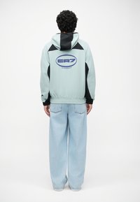 Svetlo modra kapuca jakna z črnimi detajli in elastičnim robom. Na hrbtni strani ima krogasto EA7 logo. Ohlapne svetlo modre jeans hlače.