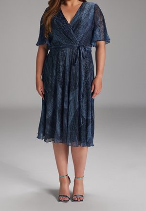 Femme portant une robe portefeuille bleu marine longueur genou avec des manches volantées, associée à des sandales à talons hauts à bride cheville turquoise sur un fond gris.