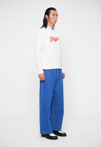 Sweatshirt branco com o logo "Fiorucci" em vermelho, combinado com calças de ganga azuis largas. O modelo usa sapatos pretos com detalhes brancos, posando contra um fundo simples.