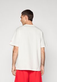 Hvid bomulds T-shirt med korte ærmer, rund halsudskæring og en lille rød detalje på venstre ærme. Modellen bærer røde shorts.
