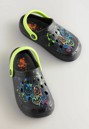 Schwarze Clogs mit grünem Akzentriemen, ausgestattet mit bunten Spritzmustern und cartoonartigen Gespenstgrafiken in Orange, Blau und Grün.