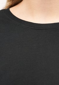 Κοντινή λήψη ενός ατόμου που φοράει απλό μαύρο μπλουζάκι με στρογγυλή λαιμόκοψη, εμφανίζοντας τον λαιμό και την άνω περιοχή του στήθους με λεπτομέρειες ριμπ στην περιφέρεια του γιακά.