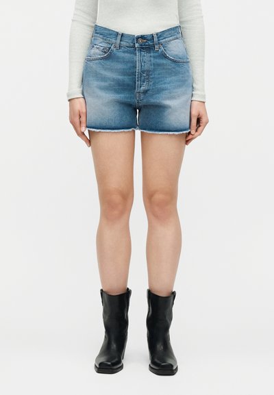 Άτομο που φοράει ανοιχτό μπλε τζιν σορτς με ξεφτισμένα τελειώματα, ανοιχτόχρωμο μακρυμάνικο τοπ και μαύρες μπότες μέχρι τη μέση της γάμπας, στέκεται μπροστά σε λευκό φόντο.