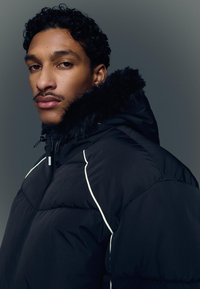 Musta toppatakki, jossa on turkisvuorattu huppu, valkoiset koristesaumat ja vetoketjusuljenta. Teksturoitu pinta ja ylisuurikokoinen malli.