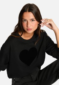 Sweatshirt noir avec un grand motif de cœur velouté noir sur le devant, col rond, et accents de boutons dorés sur l'épaule gauche.