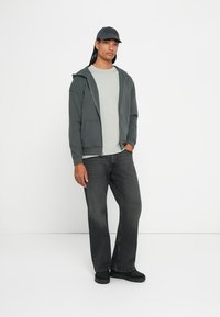 Felpa zip grigio scuro con tasche frontali, t-shirt grigio chiaro, jeans neri a zampa d'elefante e calzature nere abbinate a un cappellino coordinato.