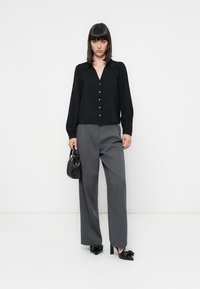 Blusa preta de botões com mangas longas, combinada com calças cinzentas largas. Bolsa preta e sapatos de salto alto com detalhe de laço completam o look.
