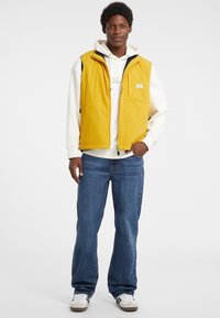 Gelbe ärmellose Weste mit einer vorderen Tasche, getragen über einem weißen Kapuzenpullover, kombiniert mit blauen Jeans und weißen Turnschuhen mit grauen Akzenten.