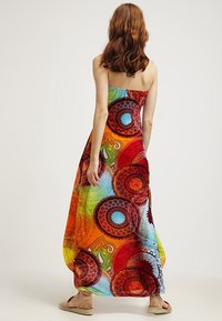 Femme aux cheveux mi-longs portant une robe maxi colorée sans bretelles avec des motifs circulaires, debout pieds nus devant un fond uni.