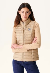 Beige Steppweste mit hohem Kragen, horizontalem Steppmuster und rundem Logo-Patch, kombiniert mit einem langärmeligen beigen Oberteil und dunklen Jeans.