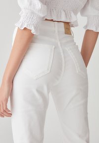 Jean blanc taille haute avec une texture lisse, comprenant deux poches arrière et un détail de couture discrète. Étiquette logo en cuir simple sur la ceinture.