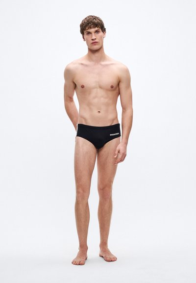 Schwarze Badehose mit tiefem Schnitt und weißem Logo an der Seite. Glattes Material und minimalistisches Design, geeignet für Badebekleidung.