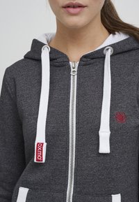 Šedá mikina na zip s bílými šňůrkami, má nášivku s logem a texturovanou tkaninu. Obsahuje přední kapsu a kovový zip.