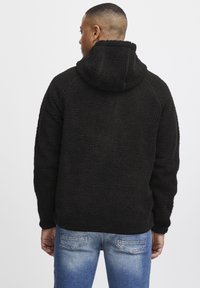 Svart fleece hettejakke med en teksturert, fluffy overflate. Har raglanermer og en avslappet passform, kombinert med blå denimjeans.