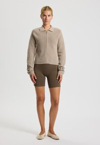 Béžový žebrovaný pletený svetr na zip s golierom, skombinovaný s hnedými šortkami a tkanými slip-on topánkami, s hladkým povrchom.