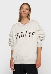 Sudadera de color beige claro con "10DAYS" impreso en marrón oscuro. Presenta un cuello redondo, hombros caídos y puños acanalados. Textura suave.