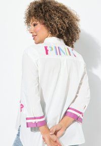 Witte blouse met lange mouwen met kleurrijke "PINK" borduring op de rug, afgewerkt met paarse en witte gestreepte manchetten. Zachte stoftextuur.