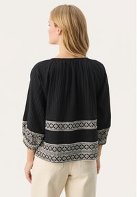 Zwarte blouse met een textuurstof, gerimpelde halslijn en lange mouwen met ingewikkelde witte borduursels langs de zoom en manchetten.