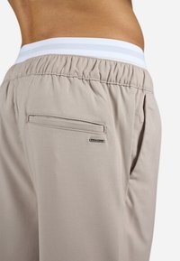 Beige bukser med elastisk talje og hvide striber, en baglomme med vandret åbning og en lille logo detalje.