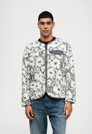 Casaco de fleece branco com padrão floral preto, colarinho redondo, fecho éclair completo, aba de bolso cinza e acabamentos pretos. Usado sobre um denim azul.