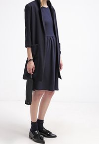 Robe bleu marine avec un haut ajusté et une jupe évasée, associée à un long cardigan bleu marine, des mocassins noirs et une pochette noire pour le poignet.