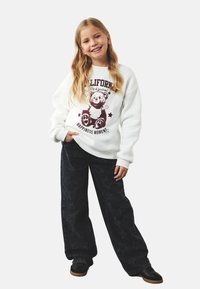 Hvid sweatshirt med teddy-bear grafik og tekst, parret med mørke denimbukser med et subtilt stjerne mønster. Sorte sko fuldender outfittet.