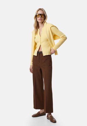 Mujer con cárdigan amarillo abotonado y gafas de sol, pantalones marrones de tiro alto y pierna ancha, y mocasines marrones, de pie con una mano en la cadera.