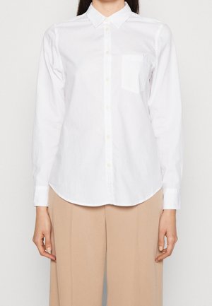 Personne portant une chemise blanche à manches longues boutonnée avec une poche poitrine, associée à un pantalon beige taille haute sur un fond uni.