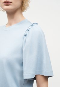 Kobieta w jasnoniebieskim topie z krótkim rękawem i falbankami na ramieniu, o kręconych blond włosach, na jednolitym białym tle.