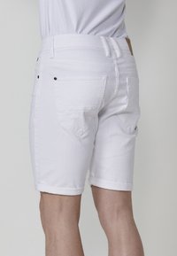 Moški v zavihanih belih denim hlačah in belem t-shirtu, prikazan zadaj proti enotni ozadju.