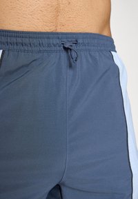 Närbild av en person som bär blåa sportshorts med justerbar dragsko och ljusblå sidor, vilket visar en del av den nedre delen av torso.