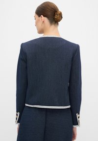 Navy sininen räätälöity takki, joka on valmistettu denimmistä, sisältäen Zikzak-reunat kauluksessa ja hihoissa, sekä kultaiset napit hihoissa.