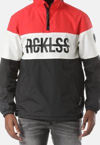 Uomo che indossa una giacca pullover rossa, bianca e nera con "RCKLSS" stampato in nero sul petto e un collo a mezza zip.