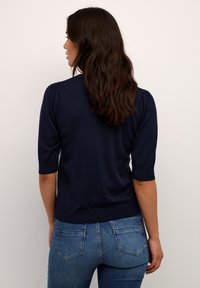 Pull en maille navy à manches courtes, avec un ourlet côtelé et des épaules légèrement bouffantes. Associé à un jean en denim bleu.