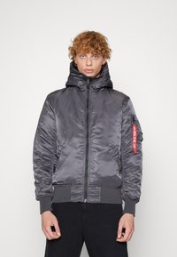Gray puffer jacket with a high collar, front zipper, side pockets, and a red tag on the sleeve. Smooth, shiny material with ribbed cuffs.